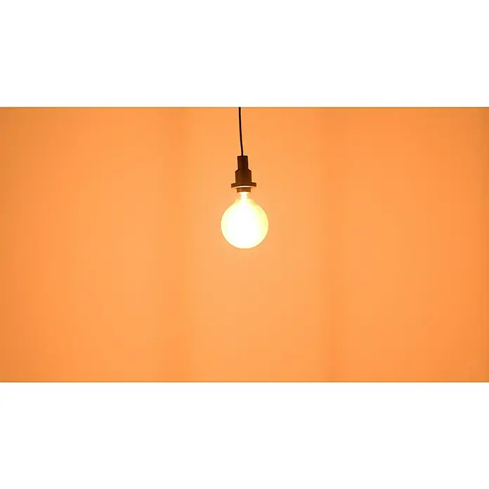 Osram LED-Lampe Vintage Edition 1906 Globe-Form E27 - Durchmesser: 12,4 cm, E27, Dimmbar, Warmweiß, 806 lm, 8,8 W - Glühbirne hängt an einem Kabel vor einem orangefarbenen Hintergrund und spendet warmes Licht.