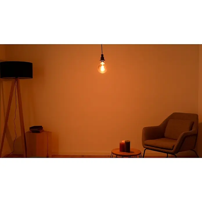 Osram LED-Lampe Vintage Edition 1906 Globe-Form E27 - Durchmesser: 12,4 cm, E27, Dimmbar, Warmweiß, 360 lm, 7,8 W - Gemütliches Wohnzimmer mit einer Glühbirne, einem Lampenschirm, einem Sessel und Kerzen, die eine warme und einladende Atmosphäre schaffen.
