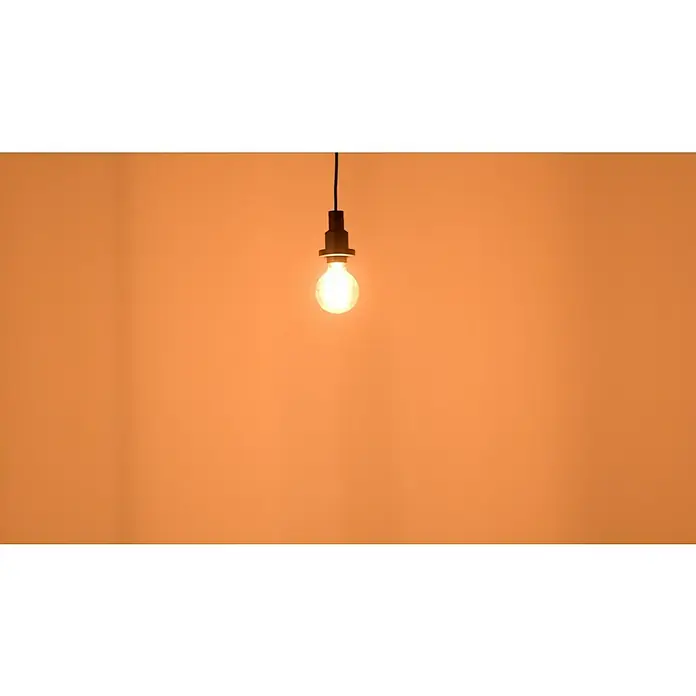 Osram LED-Lampe Vintage Edition 1906 Globe-Form E27 - E27, Dimmbar, Warmweiß, 470 lm, 5,9 W - Eine einzelne Glühbirne hängt an einem Kabel vor einem orangefarbenen Hintergrund und strahlt warmes Licht aus. Dekorative Beleuchtung im Vintage-Stil.