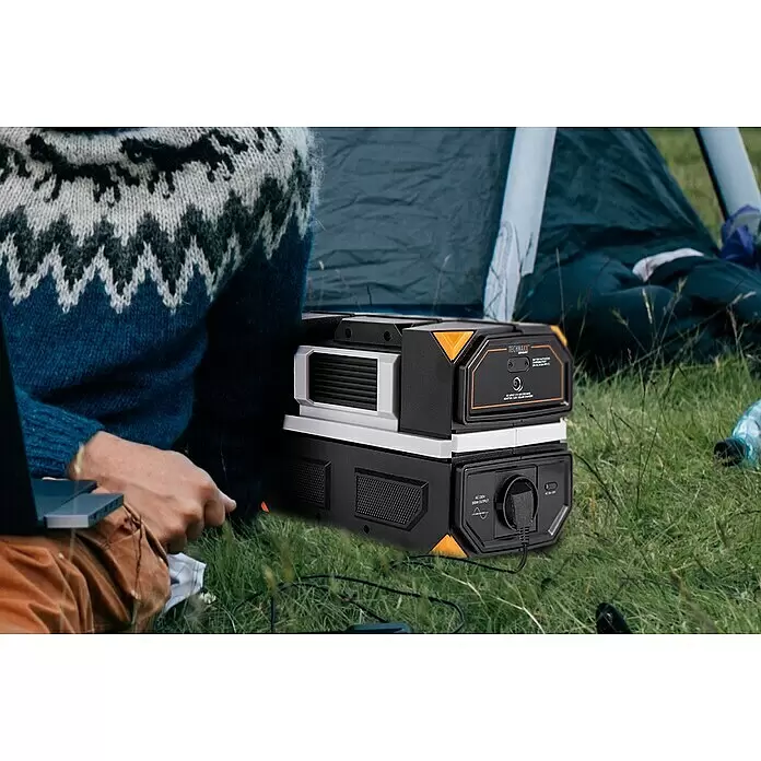 Technaxx Powerstation TX-201 - 315 Wh, Schwarz - Schwarz-weiße tragbare Powerstation mit orangefarbenen Akzenten, eingesteckt, neben einer Person beim Camping.