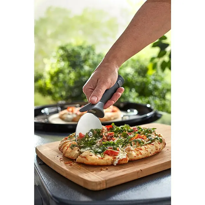 Weber Pizzaschneider - Klingendurchmesser 10 cm, Edelstahl - Nahaufnahme eines Pizzaschneiders, der eine Pizza mit frischen Zutaten auf einem Holzbrett schneidet.