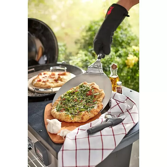Weber Pizzaschaufel - L x B: 57 x 32 cm - Eine Person in einem Ofenhandschuh verwendet eine silberne Pizzaschaufel mit schwarzem Griff, um eine Pizza vom Grill zu nehmen.