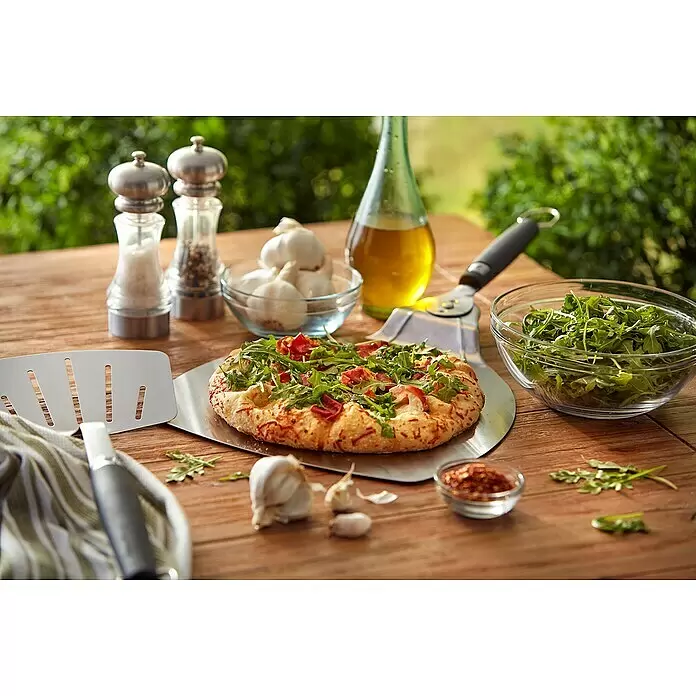 Weber Pizzaschaufel - L x B: 57 x 32 cm - Eine frische Pizza mit grünen Belägen ruht auf einer silbernen Metallpizzaschaufel, umgeben von Zutaten auf einem Holztisch.