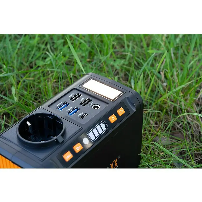 Technaxx Powerstation TX-205 - 74 Wh, Schwarz/Orange - Tragbare Powerstation mit verschiedenen Anschlüssen auf einer grünen Wiese. Ideal für Camping und Outdoor-Aktivitäten, um Geräte aufzuladen.