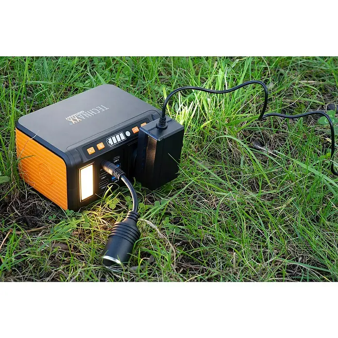 Technaxx Powerstation TX-205 - 74 Wh, Schwarz/Orange - TECHNaxx Powerstation auf Gras mit angeschlossenen Kabeln, die Vielseitigkeit und tragbare Energie demonstrieren.