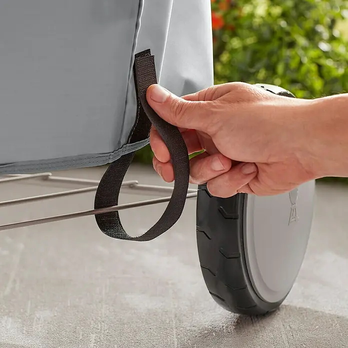 Weber Grill-Schutzhülle - Passend für: Weber Kugelgrill Ø 57 cm - Detailaufnahme einer Hand, die eine schwarze Schlaufe einer grauen Weber Grill Abdeckhaube befestigt. Schützt den Grill zuverlässig vor Wettereinflüssen.