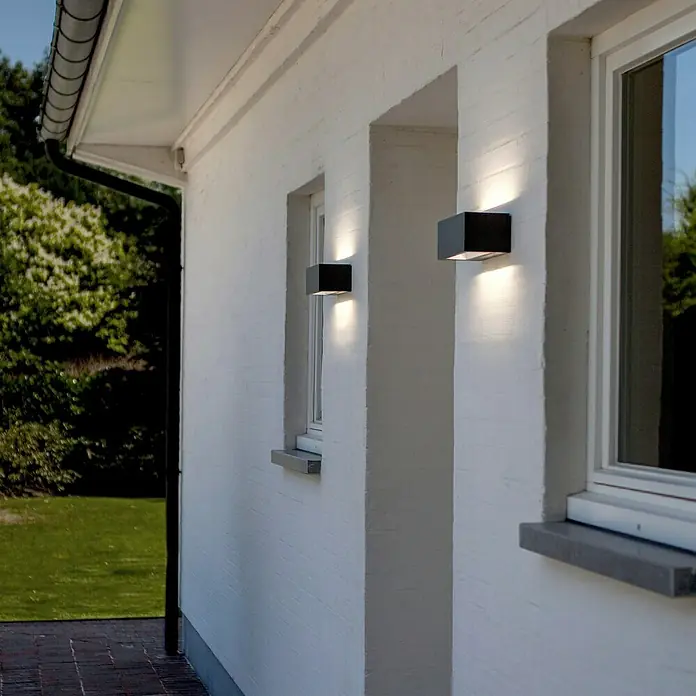 Lutec LED-Außenwandleuchte Gemini - 10,5 W, 14 x 9 x 6,5 cm, Anthrazit, IP54 - Zwei würfelförmige Wandleuchten aus schwarzem Metall strahlen helles Licht an einer weißen Hauswand aus Stein.