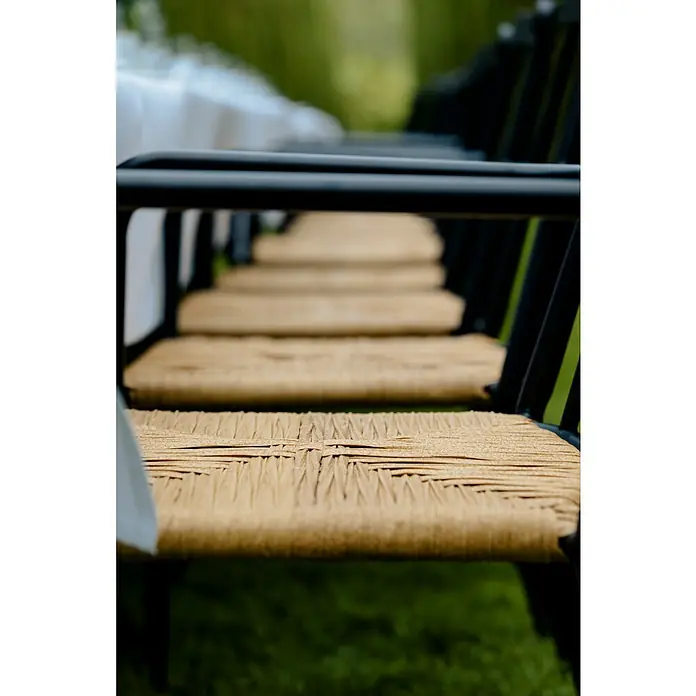 Envy Gartensessel Eva - Rattan/Geflecht, Stapelbar, Schwarz/Natur, B x T x H: 58 x 54 x 84 cm - Eine Reihe von Stühlen mit schwarzen Rahmen und gewebten, beigefarbenen Sitzen stehen an einem gedeckten Tisch für eine Veranstaltung im Freien.