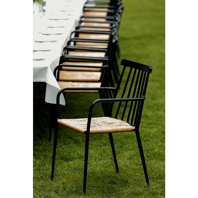 Envy Gartensessel Eva - Rattan/Geflecht, Stapelbar, Schwarz/Natur, B x T x H: 58 x 54 x 84 cm - Eine Reihe von schwarzen Stühlen mit geflochtenen Sitzen stehen neben einem Tisch mit weißer Tischdecke im Grünen.