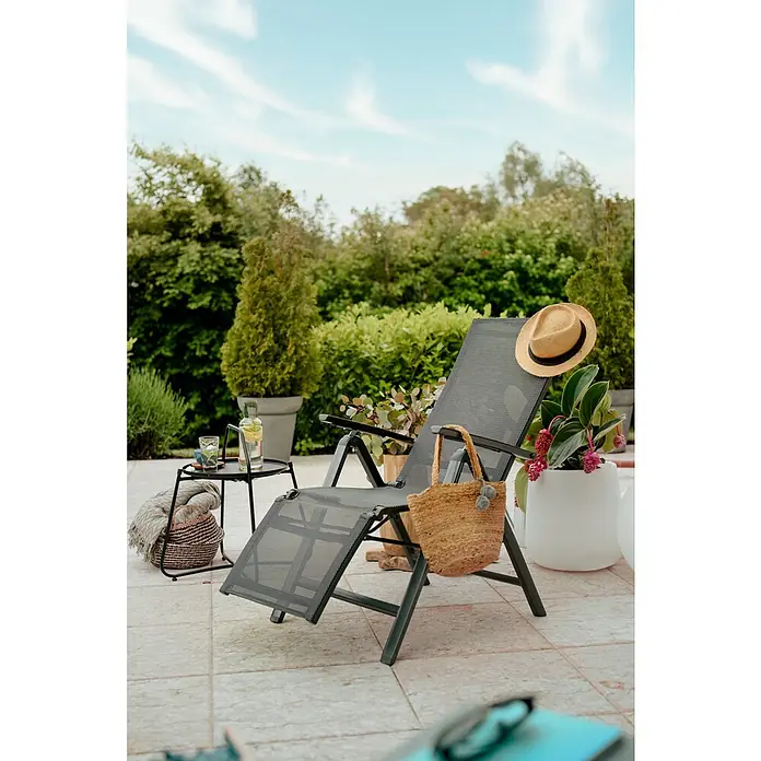 Sunfun Maja Tumbona reclinable - Aluminio/Textiline, Respaldo regulable, Antracita, An x Pr x Al: 61 x 69 x 111 cm - Una tumbona de jardín gris con un sombrero de paja y una bolsa de cesta, se encuentra en un patio con plantas y una mesa auxiliar al fondo.
