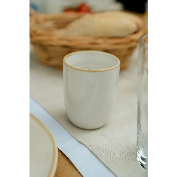 Trinkbecher TaboØ x H: 7,5 x 10 cm, Weiß, Keramik - Nahaufnahme einer eleganten, cremefarbenen Keramiktasse mit einem zarten, sandfarbenen Rand, die auf einer Tischdecke steht.