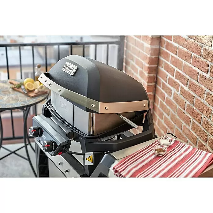 Weber Drehspieß - Länge: 16 cm, Passend für: Weber Elektrogrill Pulse 1000 / 2000 - Dunkelgrauer Weber Pulse Elektrogrill mit Edelstahlakzenten auf einem Balkon neben einer Ziegelwand und einem Tisch mit Zitronen.