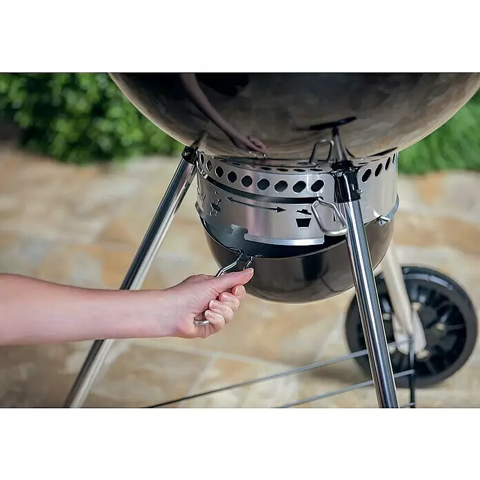 Weber Master-Touch GBS Kugelgrill Premium E-5770 - Durchmesser Grillfläche: 57 cm, Schwarz/Silber - Person verstellt die silberne Luftzufuhr eines schwarzen Kugelgrills mit Metallbeinen.