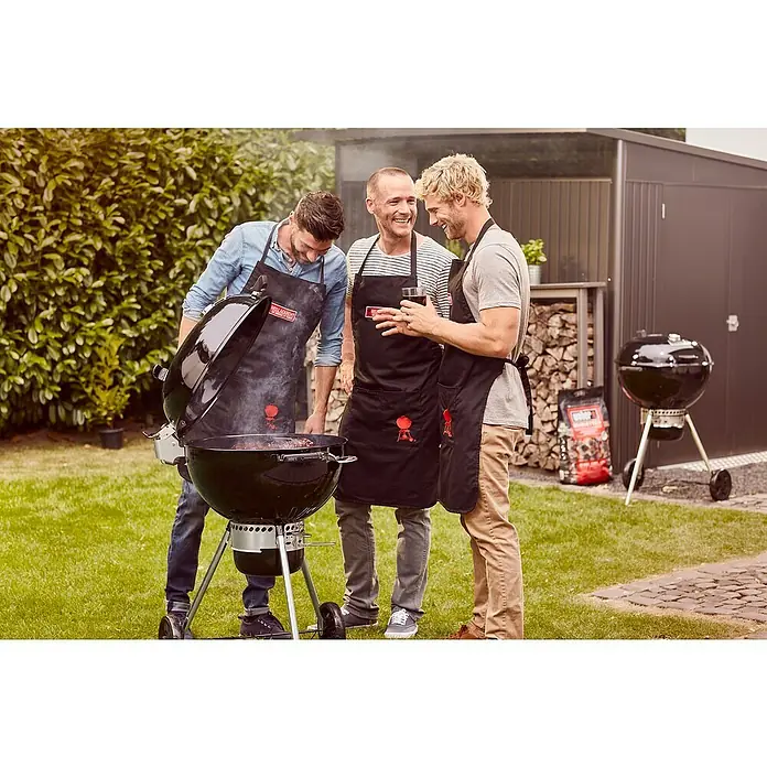 Weber Master-Touch GBS Kugelgrill Premium E-5770 - Durchmesser Grillfläche: 57 cm, Schwarz/Silber - Drei Männer stehen um einen Weber Grill und grillen Fleisch, während einer ein Getränk hält. Im Hintergrund ein Holzlager und ein Schuppen.
