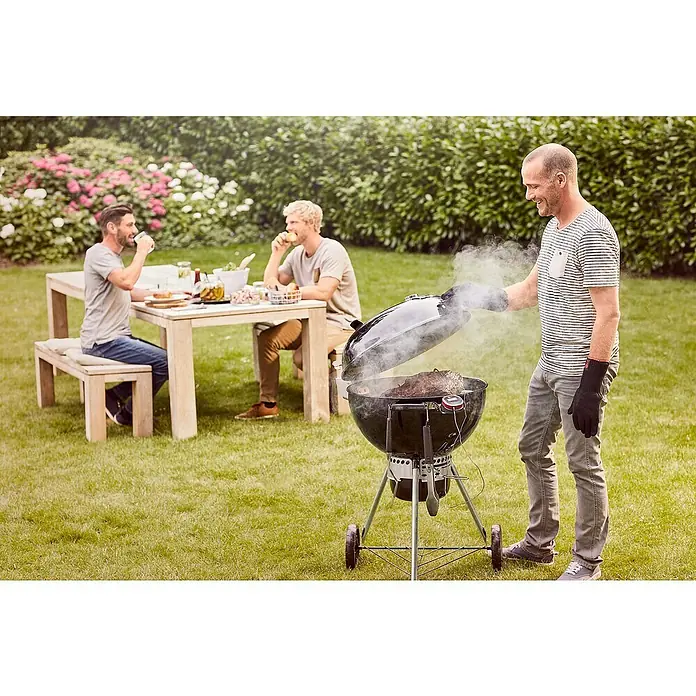 Weber Master-Touch GBS Kugelgrill Premium E-5770 - Durchmesser Grillfläche: 57 cm, Schwarz/Silber - Ein Weber-Kugelgrill wird im Garten benutzt, während zwei Männer am Tisch sitzen und essen. Der Grill erzeugt Rauch und ist mit Grillhandschuhen ausgestattet.