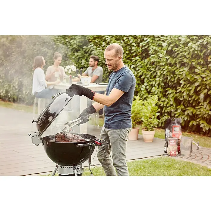 Weber Master-Touch GBS Kugelgrill Premium E-5770 - Durchmesser Grillfläche: 57 cm, Schwarz/Silber - Ein Mann grillt Fleisch auf einem schwarzen Kugelgrill im Freien, während andere am Tisch sitzen. Der Fokus liegt auf dem Weber Grill.