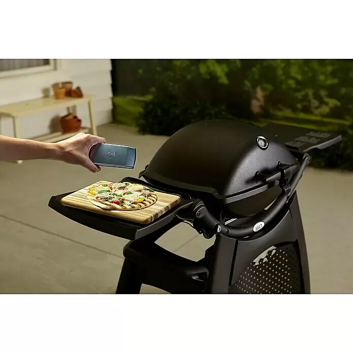 Weber Grifflicht Q - Passend für: Weber Q-Serie - Schwarzer Grill mit Pizza auf Holzbrett; eine Hand hält ein silbernes Smart-Gerät über die Pizza.