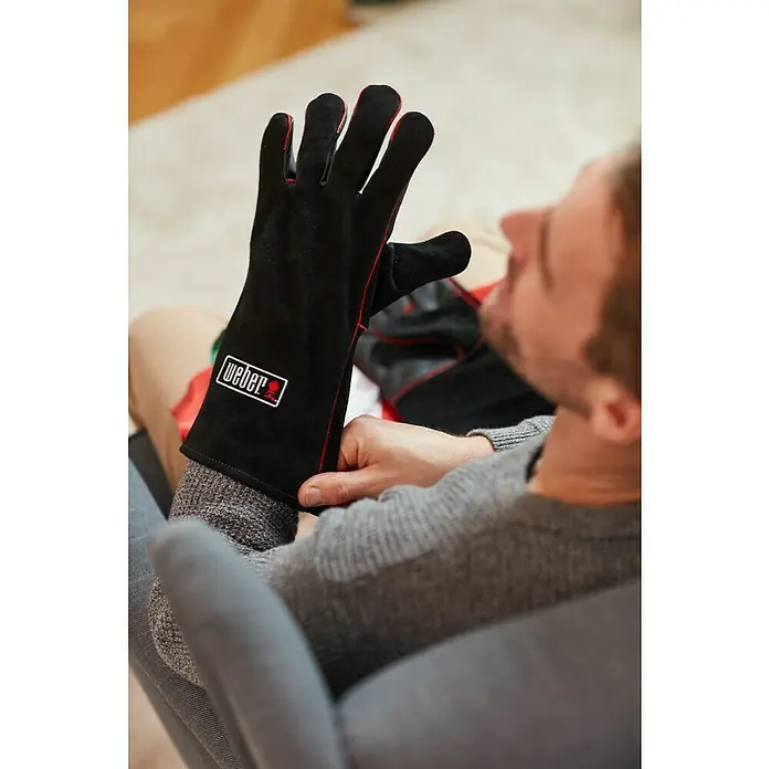 Weber Grillhandschuhe - 2 -tlg., Leder - Mann zieht einen schwarzen Weber Grillhandschuh mit roter Naht an, der das Weber Logo trägt.