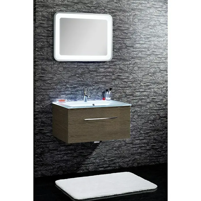 Camargue Stella Waschtischunterschrank - 59,2 x 40,8 x 39 cm, Mit Beleuchtung, Eiche Maron - Modernes Badezimmer-Interieur mit Waschtischunterschrank, Spiegel mit Beleuchtung und flauschigem, weißen Badteppich vor einer grauen Steinwand.