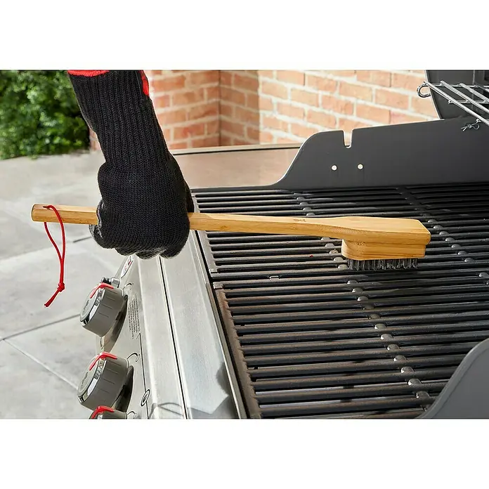 Weber Grillbürste - Länge: 44,4 cm - Nahaufnahme einer Person, die mit einer Grillbürste einen Grill reinigt. Die Grillbürste hat einen Holzgriff und Borsten aus Metalldraht.