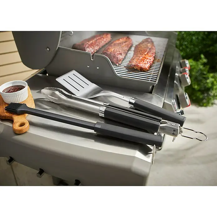 Weber Grillzubehör-Set Precision - Edelstahl - Nahaufnahme von Grillutensilien wie Zange, Spatel und Pinsel auf einem Grill, mit Fleisch auf dem Rost.