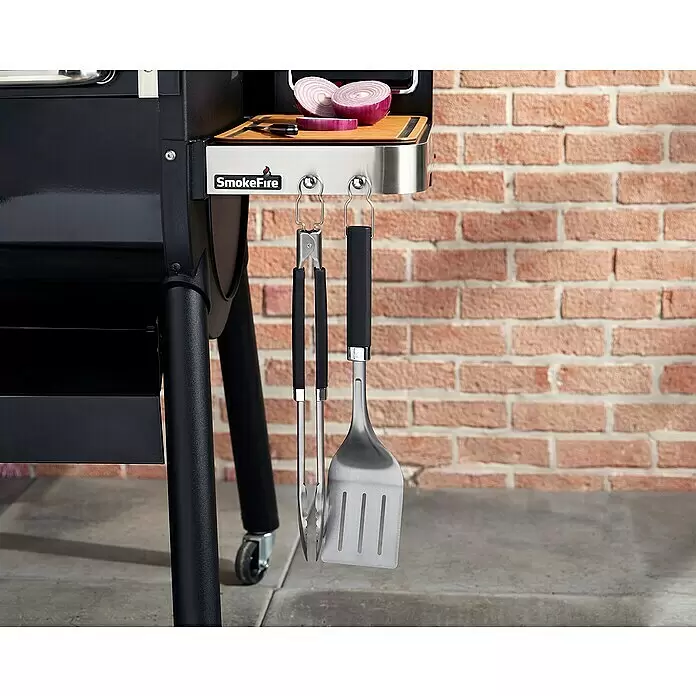 Weber Grillbesteck Set - 2 -tlg. - Schwarzer Grill mit Seitenablage, Zwiebeln und Messer. Grillzange und Spatel hängen darunter, vor einer Ziegelwand.