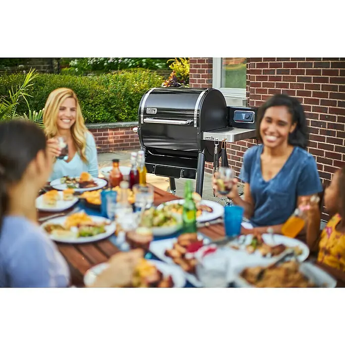Weber Pelletgrill Smokefire EX4 GBS - Grillfläche (B x T): 61 x 46 cm, Schwarz - Eine Gruppe von Freunden genießt ein Essen im Freien, mit einem Weber-Grill im Hintergrund, der für köstliche Grillgerichte sorgt.