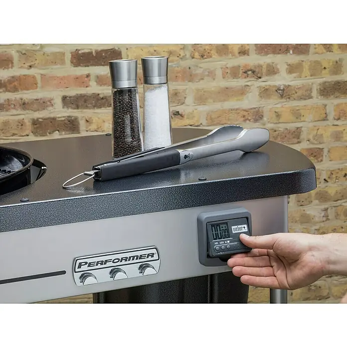 Weber Barbacoa de carbón Performer premium GBS 57 - Diámetro de la superficie de la parrilla: 57 cm, Negro - Detalle de una barbacoa Weber con temporizador, pinzas y saleros en la mesa auxiliar.