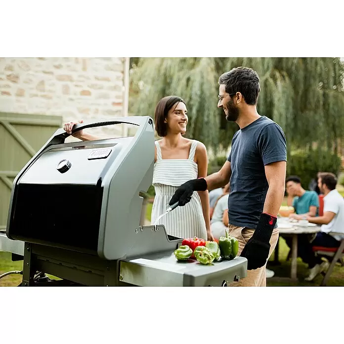 Weber Barbacoa de gas Spirit II E-310 + 7 accesorios - Superficie de la parrilla (An x Pr): 60 x 46 cm, 8,7 kW, Número de quemadores: 3 - Una pareja sonríe mientras hace una barbacoa en una parrilla negra y gris con pimientos en la mesa lateral, disfrutando de una reunión al aire libre.