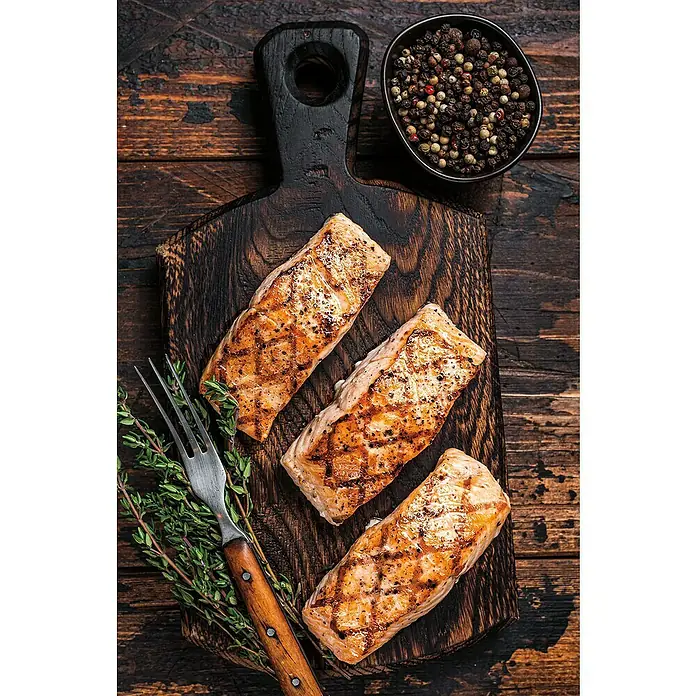Weber Crafted Grillrost Sear Grate - L x B: 41,4 x 40,6 cm, Gusseisen - Gegrillte Lachsfilets auf einem dunklen Holzbrett, garniert mit frischen Kräutern und einer Gabel, neben einer Schale mit Pfefferkörnern.