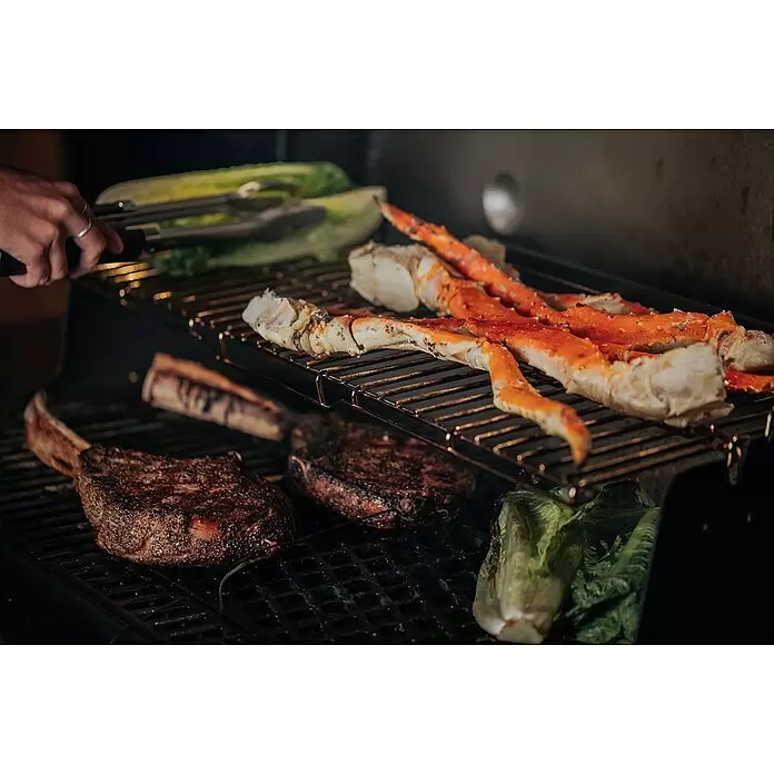 Weber Crafted Parrilla para barbacoa Sear Grate - L x An: 41,4 x 40,6 cm, Hierro fundido - Patas de cangrejo real, dos filetes grandes y lechuga asándose en una barbacoa oscura. Una mano con pinzas es visible.