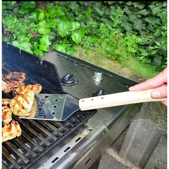 Jim Beam Grillbesteck Set - 5 -tlg. - Ein Grillspatel von Jim Beam wendet gegrilltes Hähnchen auf einem Grill im Freien, umgeben von üppigem Grün.