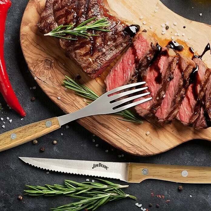 Jim Beam Steakmesser-Set II - Edelstahl, Silber/Braun, 8 -tlg. - Gezacktes Jim Beam Steakmesser und Tranchiergabel mit Holzgriffen, mit tranchiertem Steak auf Schneidebrett.