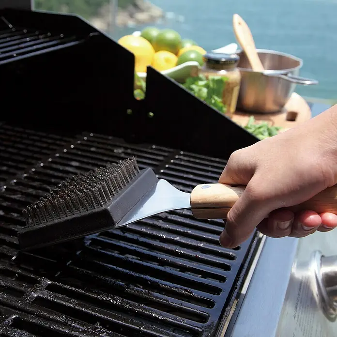 Jim Beam Grillbürste - Länge: 46 cm - Eine Grillbürste mit Drahtborsten wird verwendet, um einen Grillrost zu reinigen. Im Hintergrund ist Essen zu sehen.