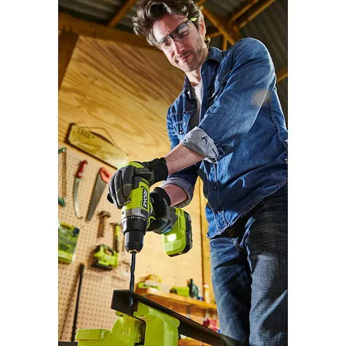 Ryobi ONE+ Akku-Schlagbohrschrauber RPD18-0 - 18 V, Ohne Akku, 52 Nm - Ein Mann bohrt mit einem Ryobi-Bohrer durch Metall, das in einem Schraubstock befestigt ist. Er trägt eine Schutzbrille und Handschuhe.
