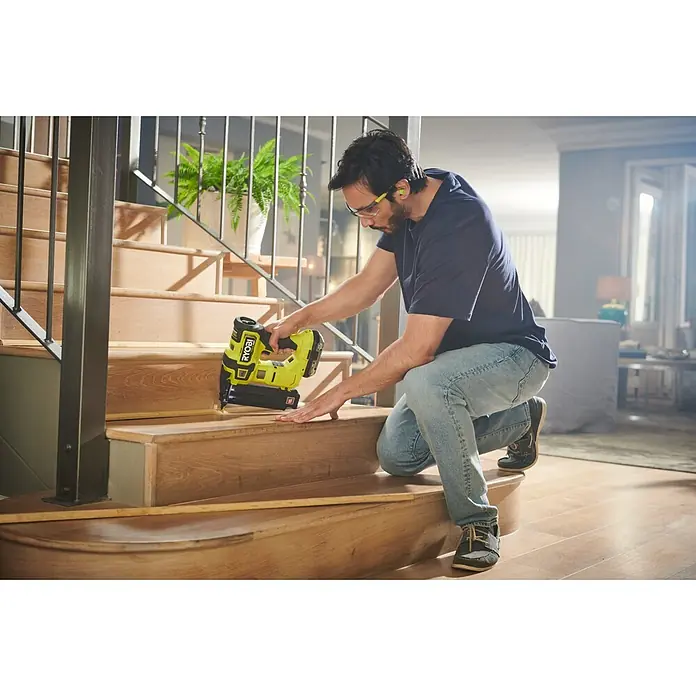 Ryobi ONE+ Akku-Nagler R18GN18X-0 - 18 V, Ohne Akku, Länge Nagel: 15 mm - 54 mm - Ein Mann kniet auf einer Treppe und verwendet einen gelben RYOBI-Nagelspistole, um die Treppe zu renovieren. Er trägt Schutzbrille und Ohrstöpsel.