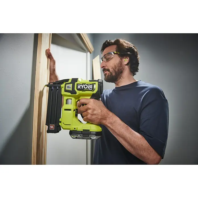 Ryobi ONE+ Maschinenpaket - Akku-Nagler R18GN18X-0 + gratis Starter-Kit - Ein Mann verwendet eine Ryobi Akku-Nagelpistole, um Holzleisten an einer Wand zu befestigen. Er trägt eine Schutzbrille und Gehörschutz.