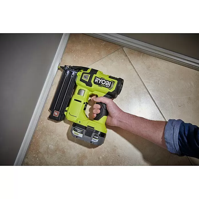 Ryobi ONE+ Akku-Nagler R18GN18X-0 - 18 V, Ohne Akku, Länge Nagel: 15 mm - 54 mm - Eine Hand hält ein grün-schwarzes Ryobi Akku-Nagelgerät an einer hellen Fußleiste und Fliesenboden, bereit zum Gebrauch.