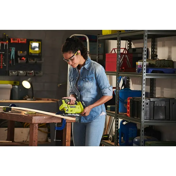 Ryobi ONE+ Akku-Pendelhubstichsäge RJS18-0 - 18 V, Ohne Akku - Eine Frau benutzt eine RYOBI Stichsäge, um ein Stück Holz auf einer Werkbank zu schneiden, wobei sie Schutzbrille und Ohrstöpsel trägt.