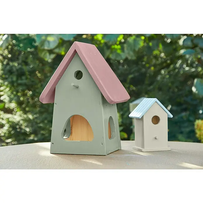swingcolor Farblasur Universal-Gartenfarbe375 ml, Seidenweiß, Seidenmatt - Zwei charmante Vogelhäuschen, das größere in Grün und Rosa und das kleinere in Weiß und Blau, stehen auf einem Tisch vor grünem Laub.