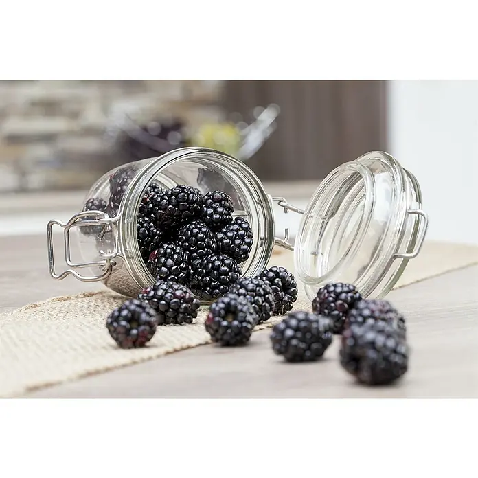 Brombeere - Rubus fruticosus 'Triple Crown', Topfvolumen: 2 l - Einmachglas mit frischen Brombeeren, teilweise verschüttet, auf einem Tisch mit rustikalem Stoffläufer. Die Beeren sind saftig und dunkel.