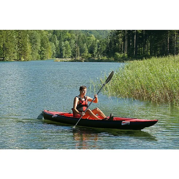 Grabner Kajak Tramper - L x B: 400 x 90 cm, Nutzlast: 200 kg, Geeignet für: 2 Personen - Ein Mann paddelt mit einem schwarzen und roten Grabner-Kajak auf einem ruhigen See, umgeben von grünen Bäumen und Schilfgras.