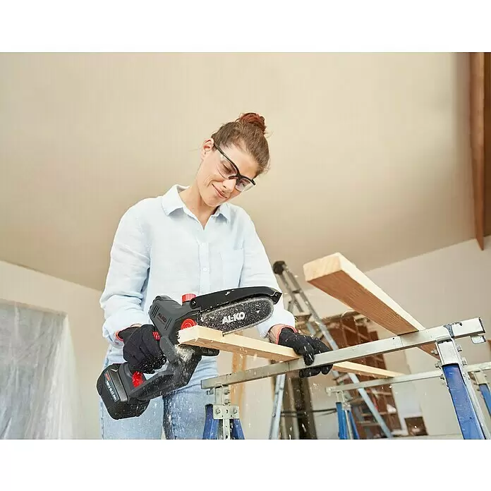 AL-KO Bosch Home & Garden Compatible Akku-Kettensäge CSM 1815 - 18 V, Li-Ionen, 1 Akku, Schwertlänge: 15 cm - Eine Frau mit Schutzbrille und Handschuhen sägt mit einer schwarz-roten AL-KO Mini-Kettensäge ein Holzbrett auf Sägeböcken.