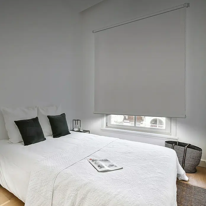 Estor enrollable opaco PP - An x Al: 150 x 180 cm, Blanco - Una persiana enrollable gris cubre la ventana sobre una cama blanca con almohadas oscuras, creando una estancia tranquila y oscura.
