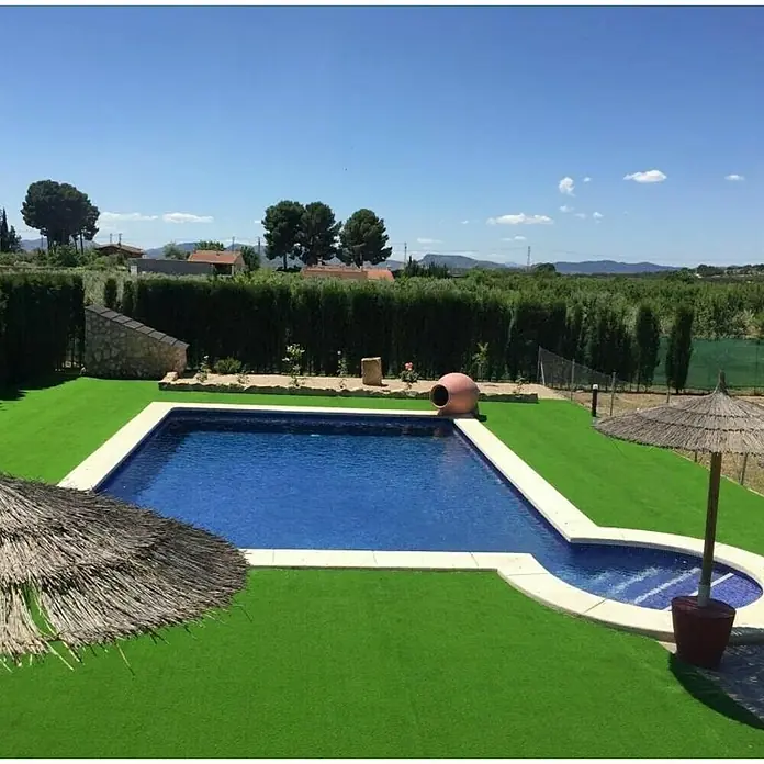 Césped artificial Habana - 5 x 2 m, Altura del pelo: 33 mm, 27.300 puntadas/m² - Atractiva zona de piscina con agua turquesa y piedras claras, rodeada de exuberante vegetación y sombrillas.