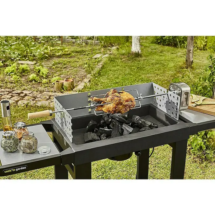 Barbacoa de carbón Easy 60 - Superficie de la parrilla (An x Pr): 57 x 30 cm, Negro - Primer plano de una parrilla de carbón con un asador que tiene un pollo dorado, rodeado de exuberante vegetación.