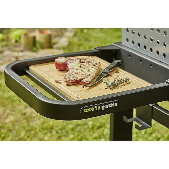 Barbacoa de carbón Easy 60 - Superficie de la parrilla (An x Pr): 57 x 30 cm, Negro - Primer plano de una parrilla con un chuletón, hierbas y un cuchillo sobre una tabla de cortar.