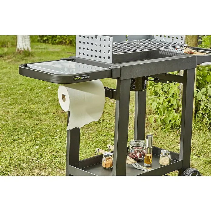 Barbacoa de carbón Easy 60 - Superficie de la parrilla (An x Pr): 57 x 30 cm, Negro - Primer plano de una parrilla "Cook In Garden" con un portarrollos de papel, especias y utensilios para asar en el estante inferior.