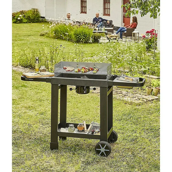 Barbacoa de carbón Easy 60 - Superficie de la parrilla (An x Pr): 57 x 30 cm, Negro - Parrilla con comida asándose en la superficie, equipada con superficies de almacenamiento, en el jardín, mientras la gente se relaja al fondo.