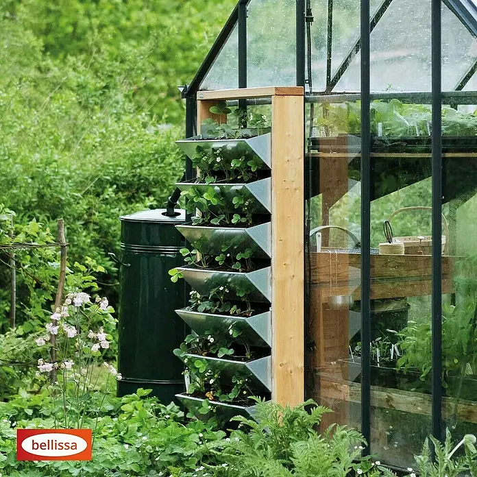 Bellissa Hochbeet Inalto L8 - 100 x 32 x 180 cm, Stahl, Hellbraun/Silber - Ein vertikaler Pflanzturm mit Erdbeerpflanzen neben einem Gewächshaus, ideal für kleine Gärten und Balkone.
